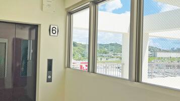 A room with a large window, a door with a number '6' sign, and a view of a building and greenery outside.