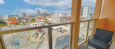 A balcony with wicker furniture, a view of a cityscape with buildings and a parking lot, and a clear sky.