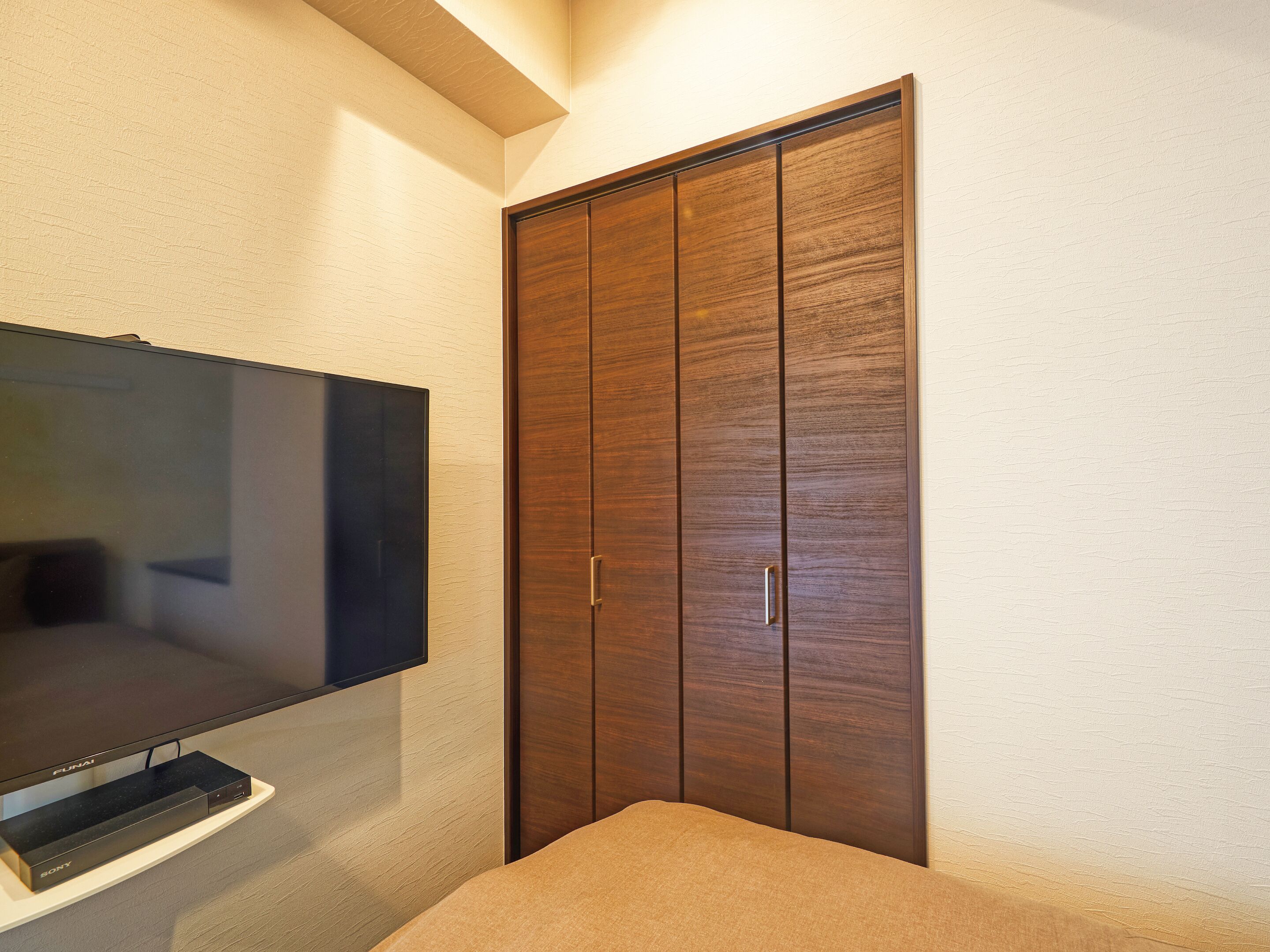 A bedroom with a TV mounted on the wall, a wooden wardrobe, and a bed with a beige blanket.