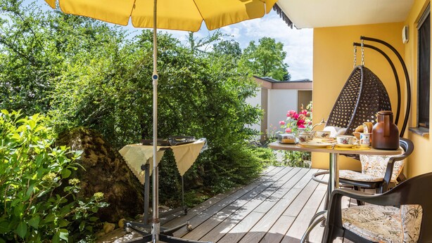 Une terrasse avec un parasol jaune, une table avec des chaises et un fauteuil suspendu.