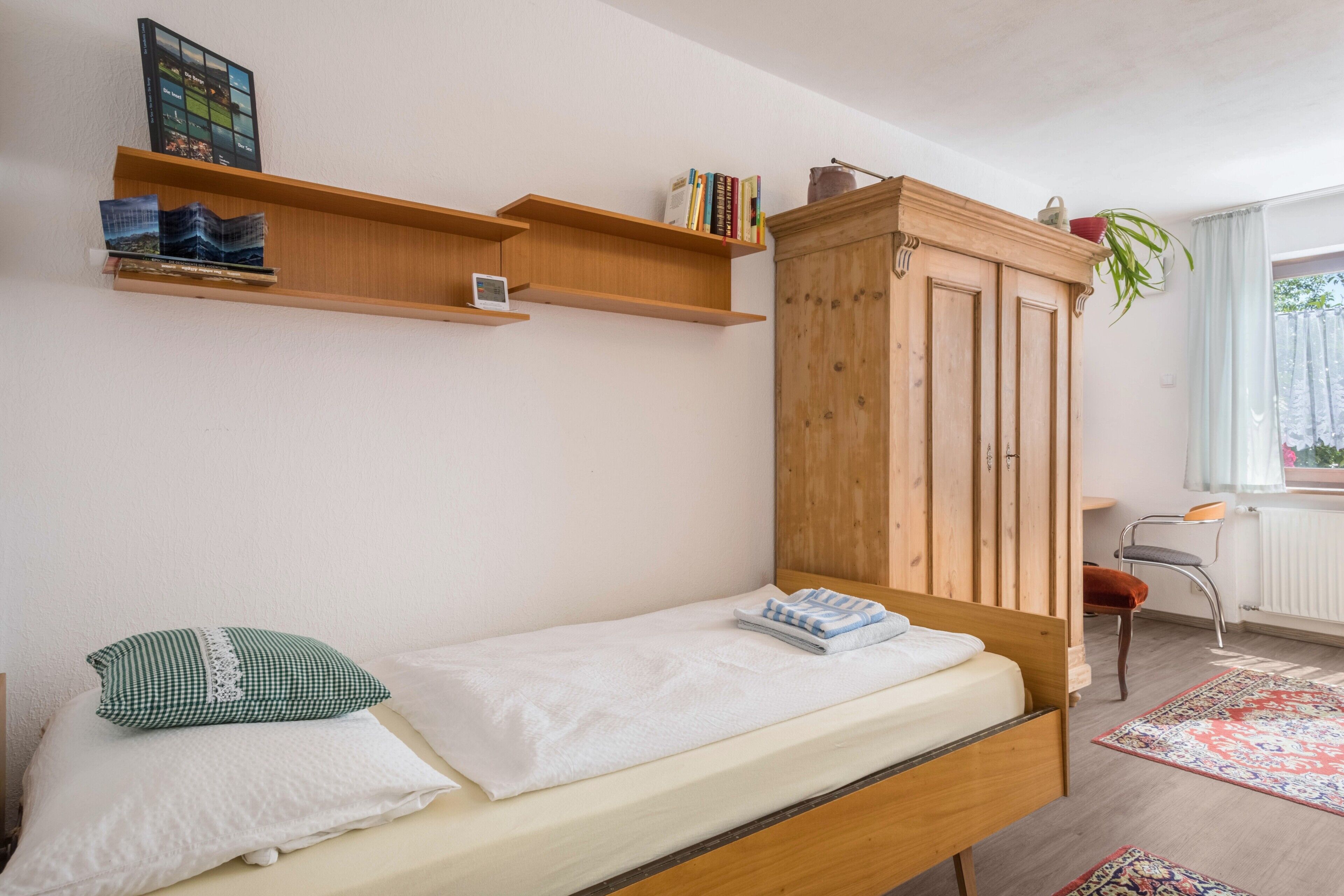 A bedroom with a single bed, a wooden wardrobe, a shelf with books, a small table, and a chair.