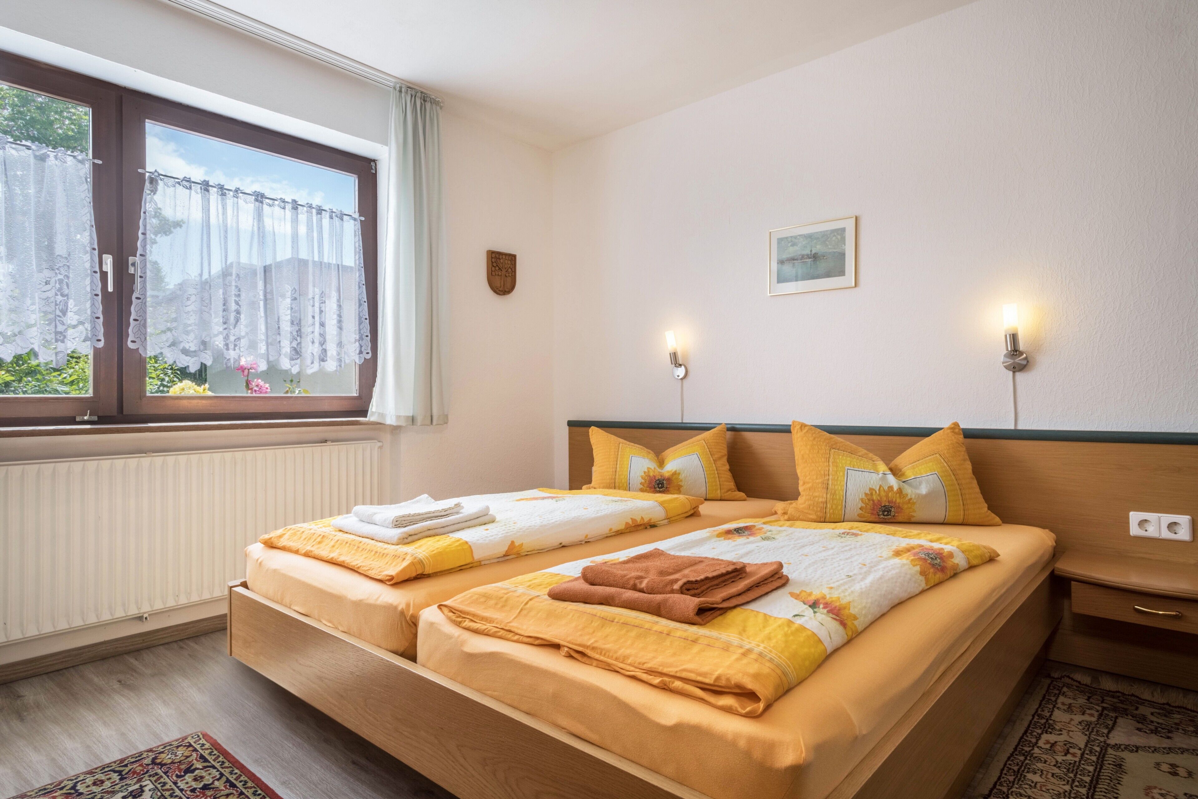 A double bed with a floral bedspread, two pillows, a nightstand, a wall-mounted light, a window with curtains, a radiator, and a small picture on the wall.
