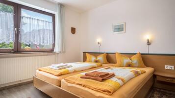 A double bed with a floral bedspread, two pillows, a nightstand, a wall-mounted light, a window with curtains, a radiator, and a small picture on the wall.