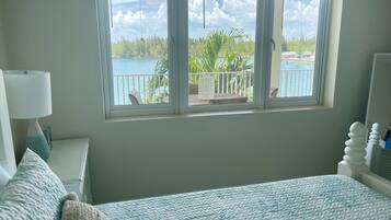 A bedroom with a bed, a nightstand, a lamp, and a window with a view of a balcony and water.