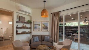 A living room with a sofa, two armchairs, a round coffee table, and a hanging light fixture. There are framed pictures on the wall, a window with curtains, and a sliding glass door leading to an outdoor area. - Capri Coastal Tuscan Getaway – Ideal for Extended Stays! (Encinitas)