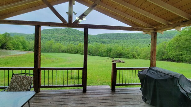 A covered outdoor deck with a view of a green field and forested hills. - Seek your rest at “Bertie’s Nest” (Parthenon)