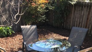 Een ronde glazen tafel met vier stoelen, op een betonnen terras met een houten hek en bomen op de achtergrond.