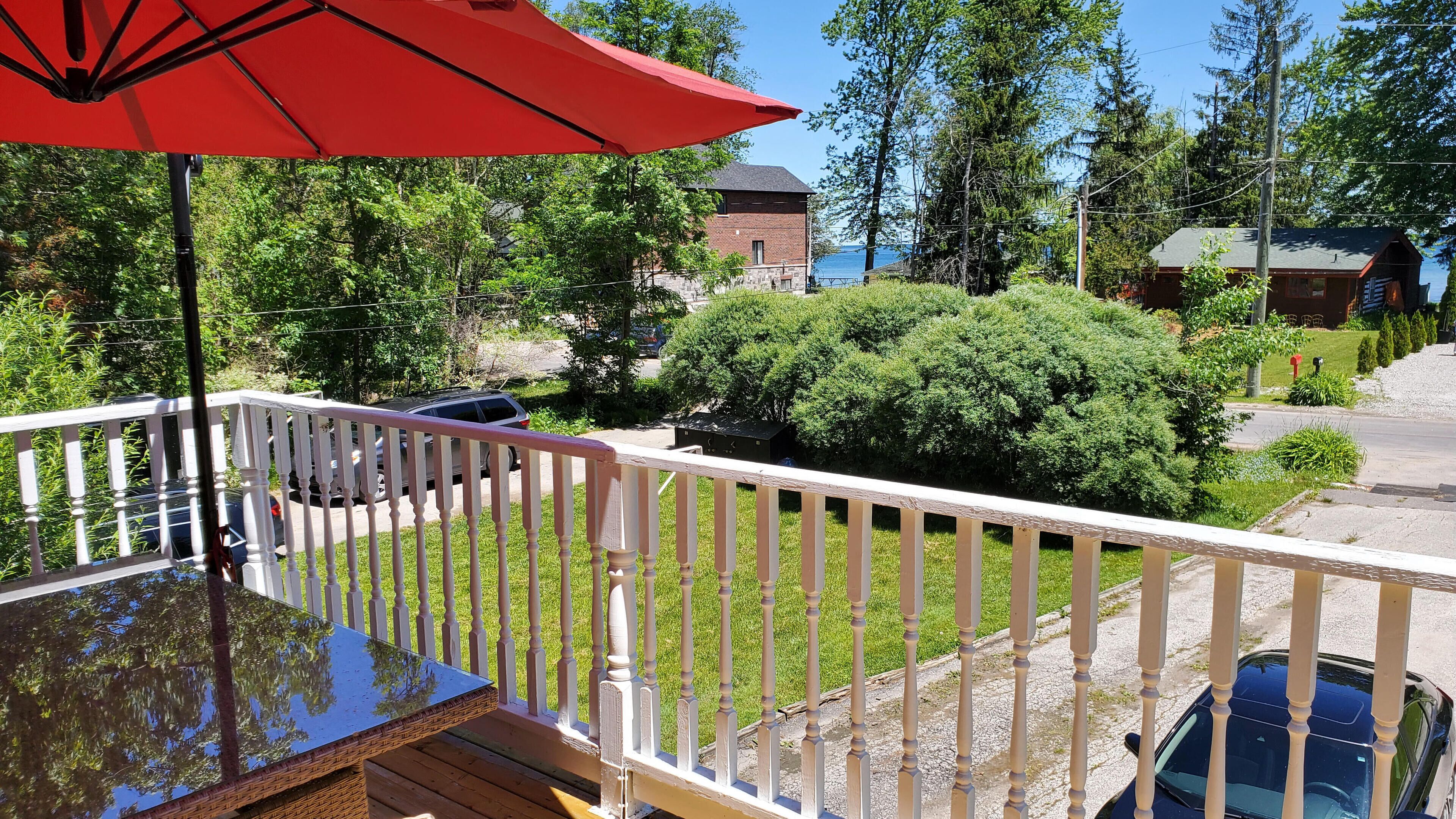 A deck with a glass table and a red umbrella, overlooking a grassy area with a car and a brick building.
