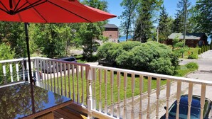 Une terrasse avec une table en verre et un parasol rouge, donnant sur un espace verdoyant où se trouvent une voiture et un bâtiment en briques.