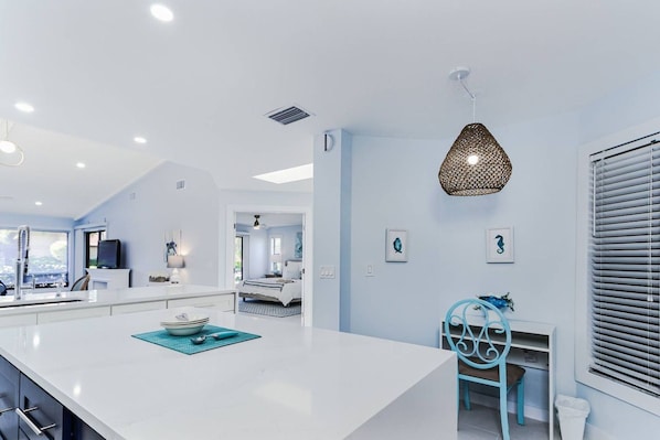 A modern kitchen with a central island, a dining area with a blue chair, and a bedroom visible through an open door. - Beach Paradise, Pool/Spa, Bright Updated Condo (Sarasota)