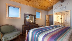A bedroom with a wooden ceiling, two windows, a bed with a colorful striped blanket, a wooden dresser, a green armchair, and a mirror.
