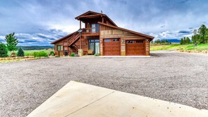 A two-story log cabin with a large garage, situated on a gravel driveway with a concrete patio in the foreground.