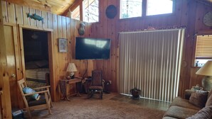 Interior acogedor de una cabaña con paredes de madera, un televisor montado en la pared, una zona de estar confortable con un sofá y sillones, y un ventanal con persianas.