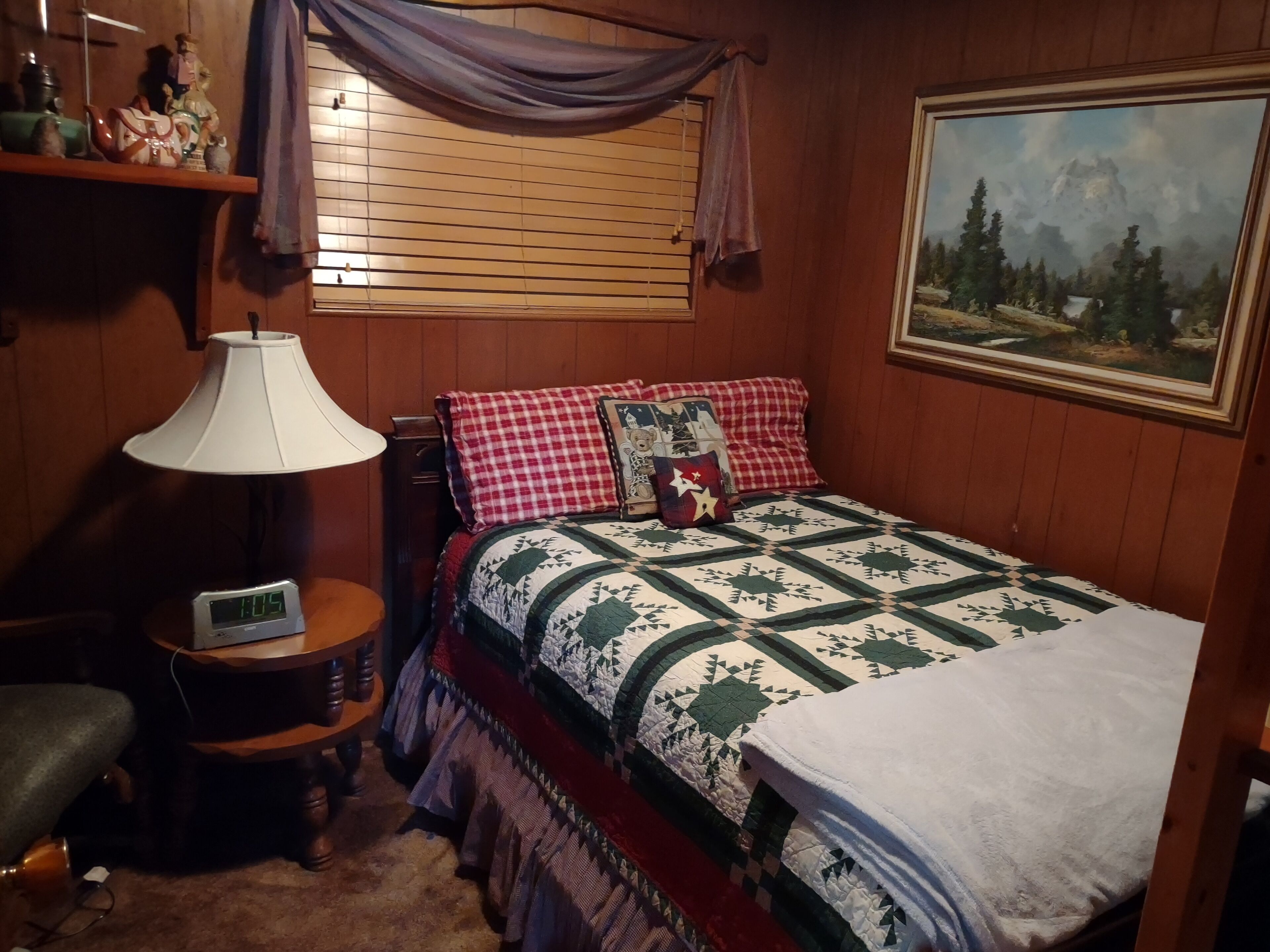 A cozy bedroom with a wooden headboard, a bed with a quilt, a bedside table with a lamp, a clock, and a painting on the wall.