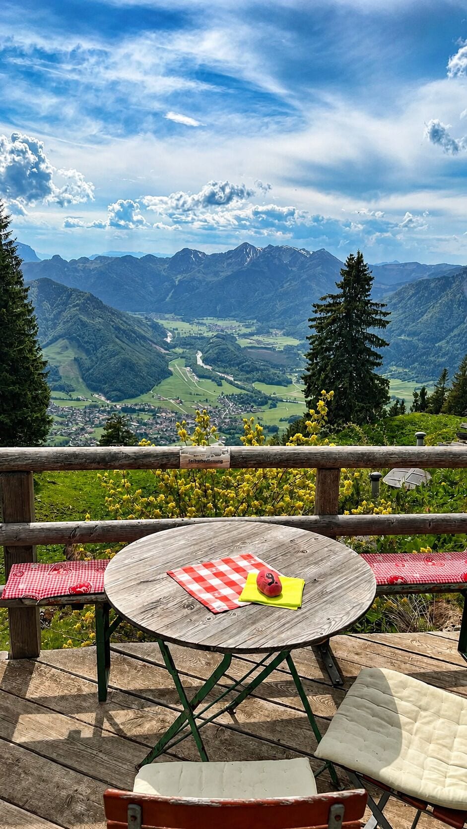 A wooden table with a checkered tablecloth and a red apple, set on a deck overlooking a mountainous landscape.