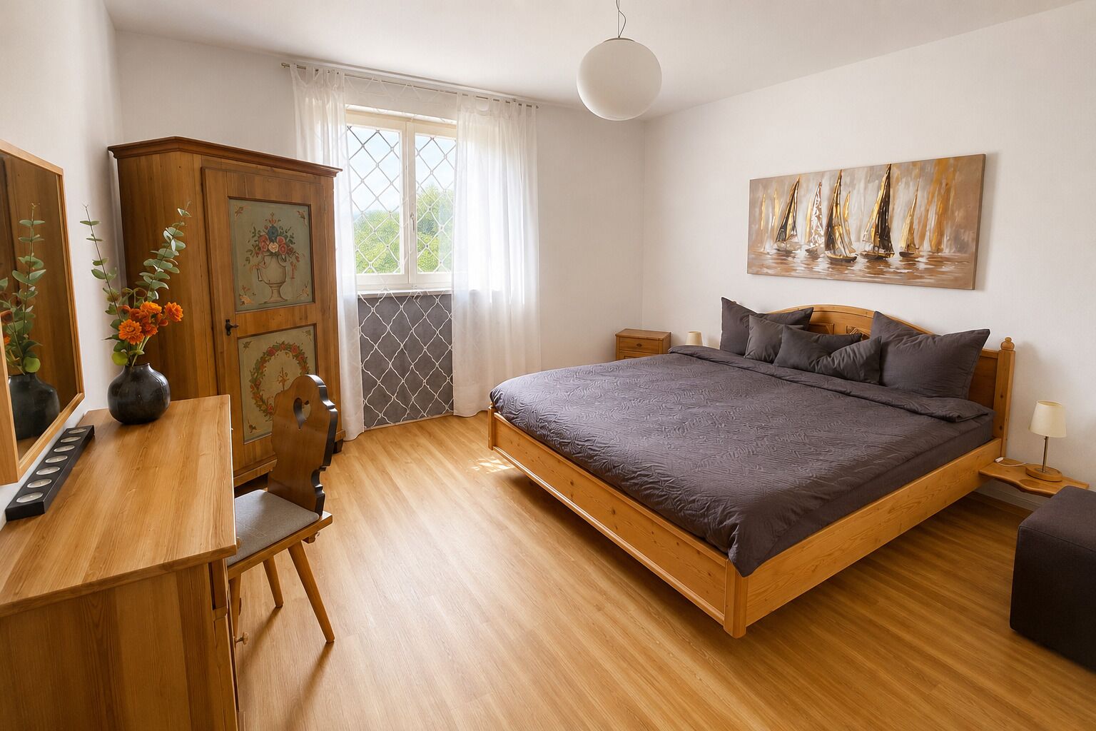 Une chambre à coucher avec un lit en bois, un bureau agrémenté d’un vase de fleurs, une armoire où sont exposées des œuvres d’art et une fenêtre avec des rideaux.