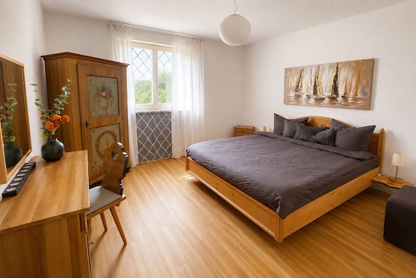 A bedroom with a wooden bed, a desk with a vase of flowers, a cabinet with artwork, and a window with curtains.