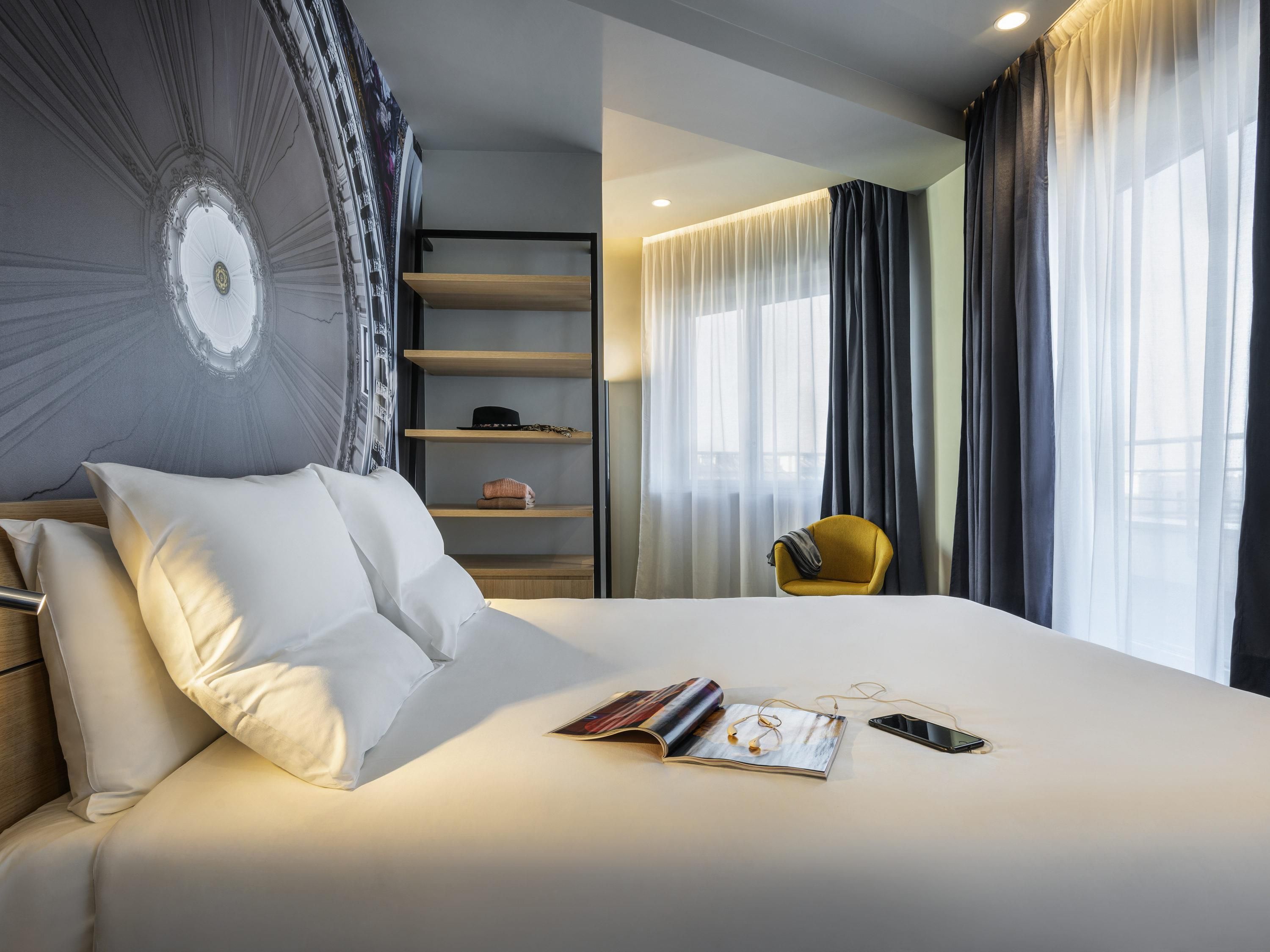 Habitación de hotel con una cama grande, una mesita de noche, una silla, una estantería y una ventana con cortinas.