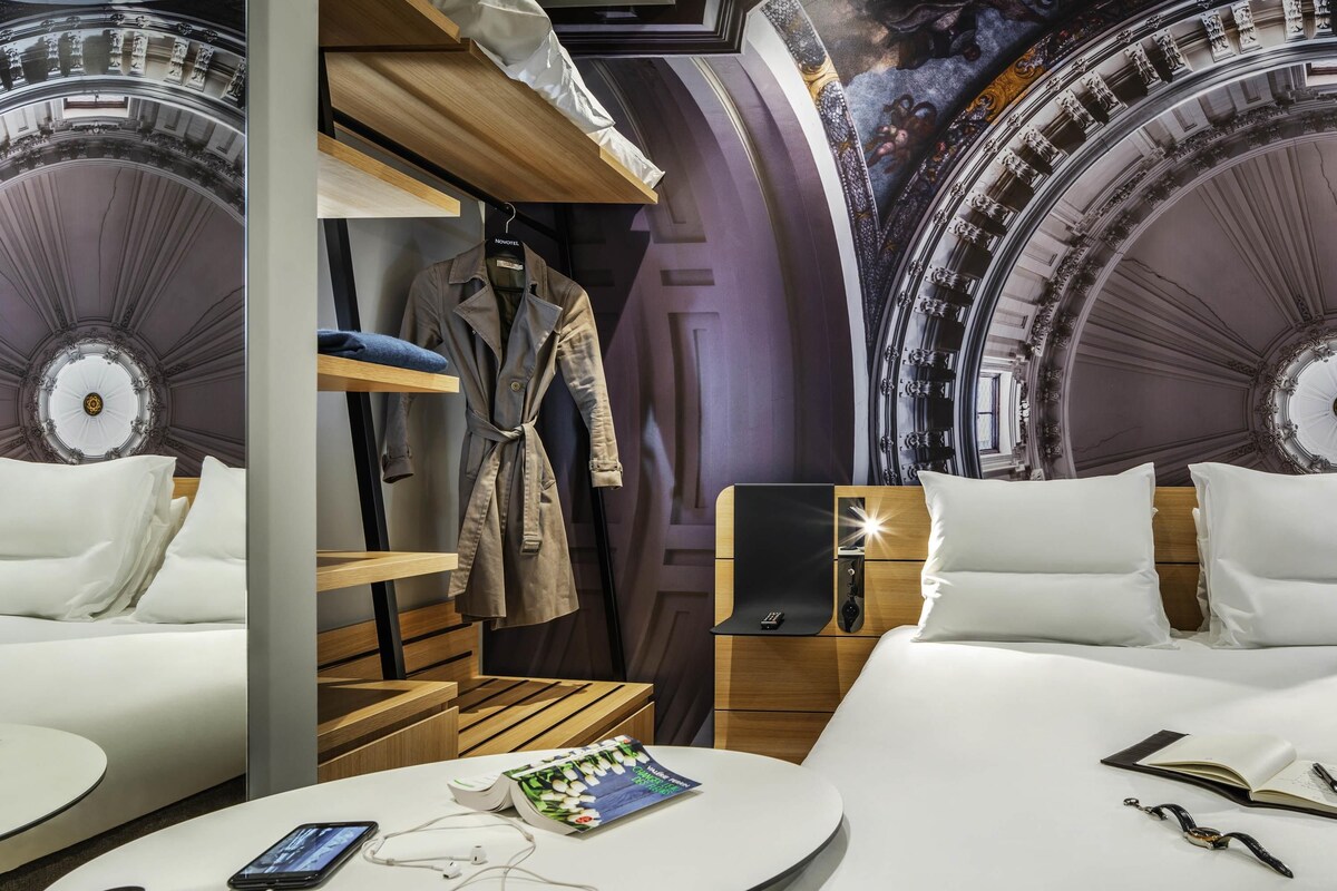 A hotel room with a bed, bedside tables, a wardrobe, a round table with a book and a phone, and a wall with a decorative ceiling.