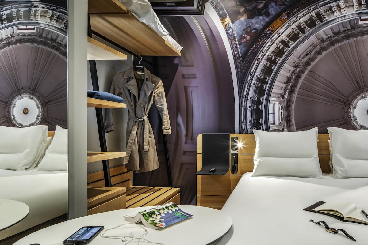 A hotel room with a bed, bedside tables, a wardrobe, a round table with a book and a phone, and a wall with a decorative ceiling.