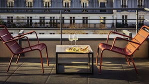 A terrace with two red chairs, a table with a glass of wine, and a view of buildings. - Novotel Belleville Paris 20 (Paris)