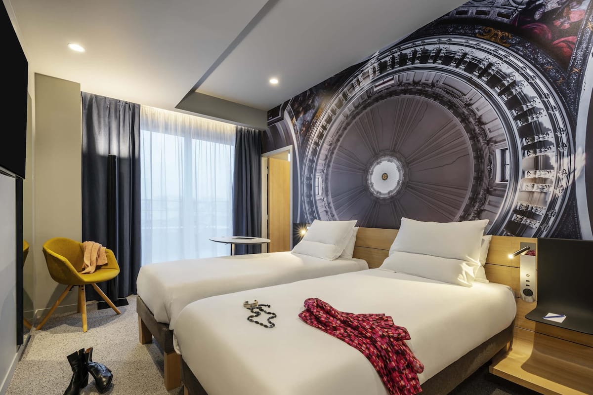 A hotel room with two beds, a small table, a chair, and a large wall mural.