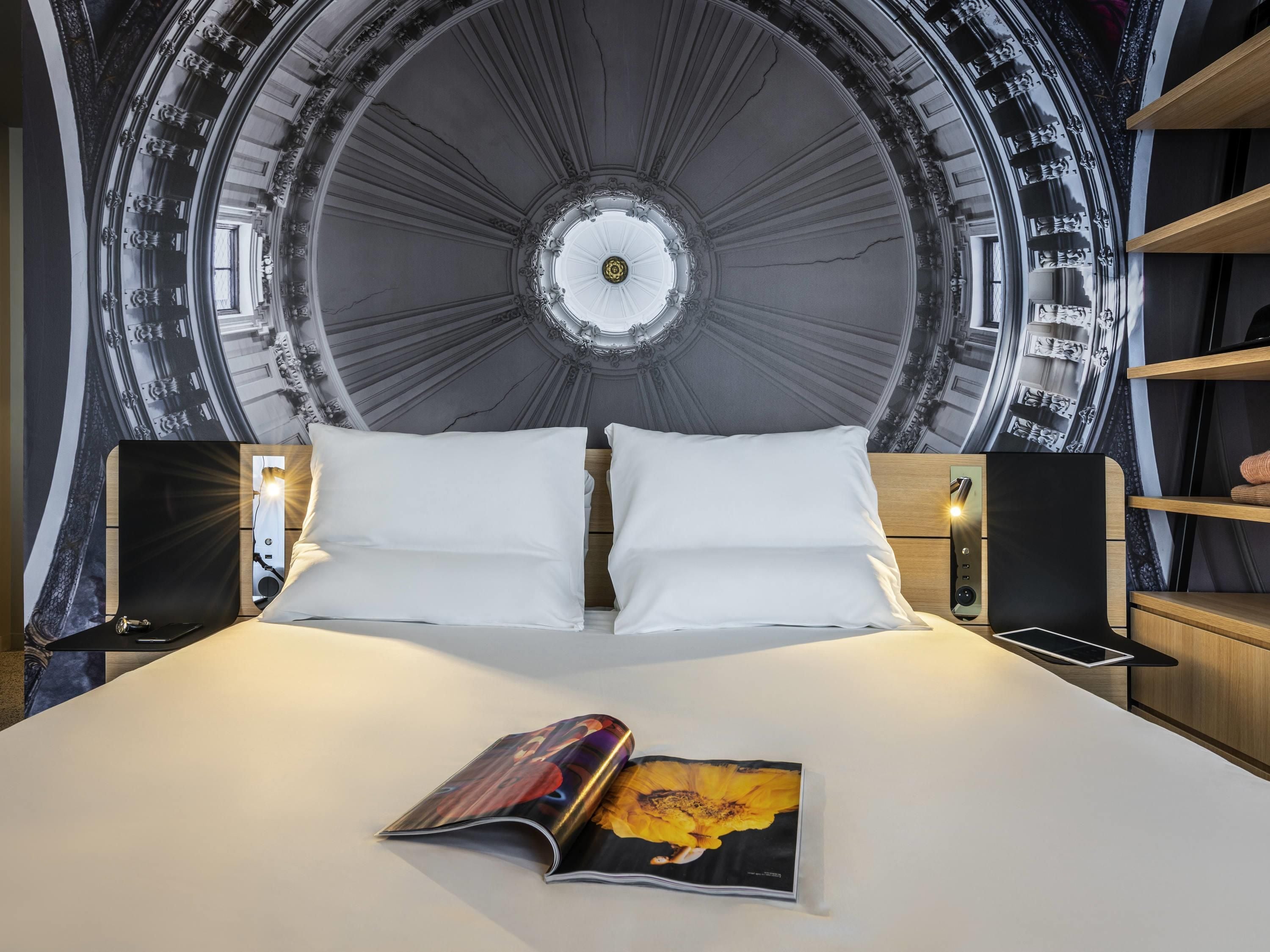 A bed with white linens, a magazine on the bed, and a large circular wall mural.
