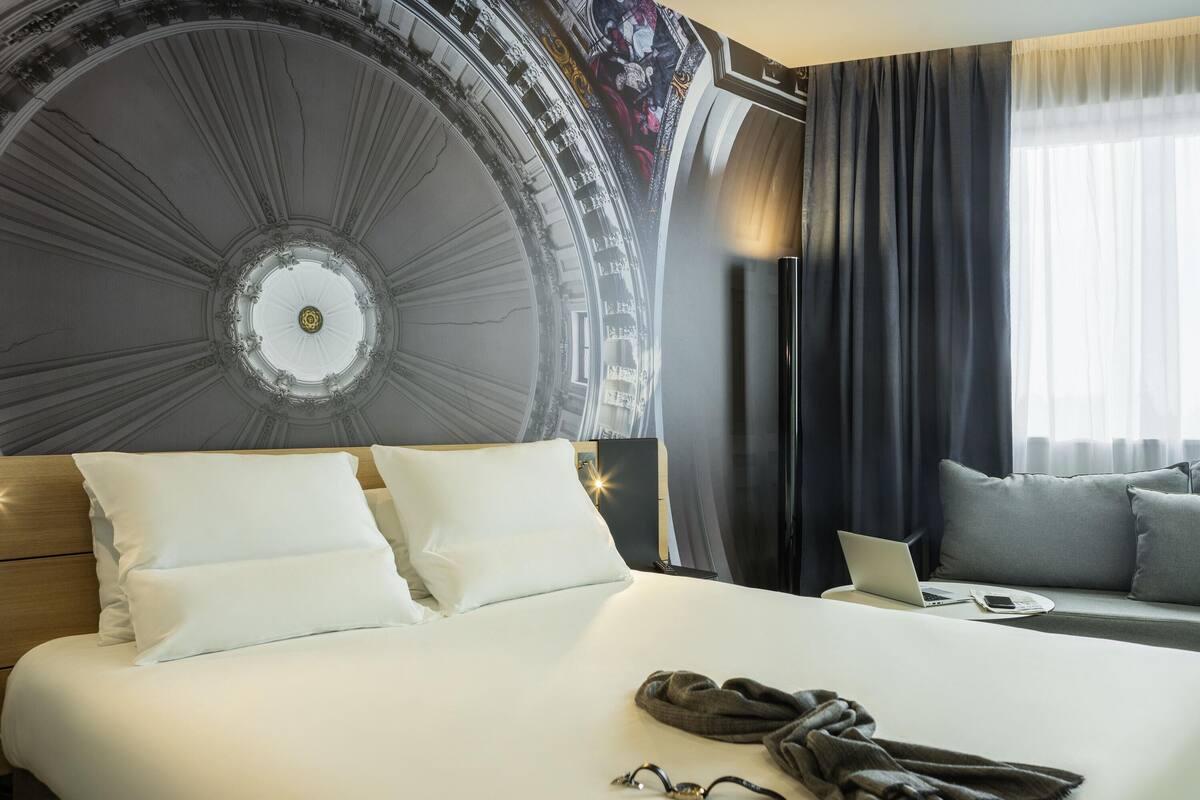 A hotel room with a large bed, a laptop on a side table, and a view of a building's facade through the window.