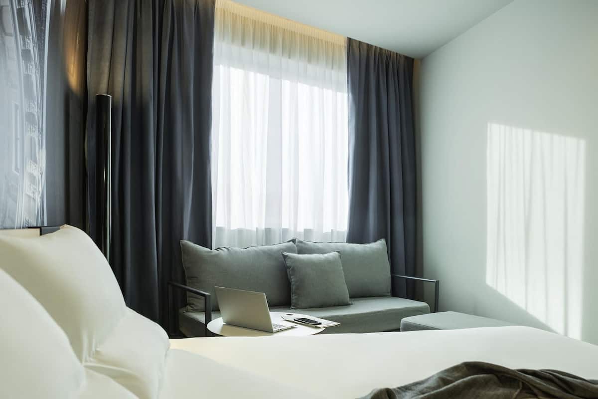 Une chambre dâhĂŽtel moderne Ă©quipĂ©e dâun lit, dâun bureau avec un ordinateur portable, dâun canapĂ© et dâune fenĂȘtre avec des rideaux.