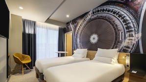 A hotel room with two beds, a small table, a chair, and a large mural on the wall. - Novotel Belleville Paris 20 (Paris)