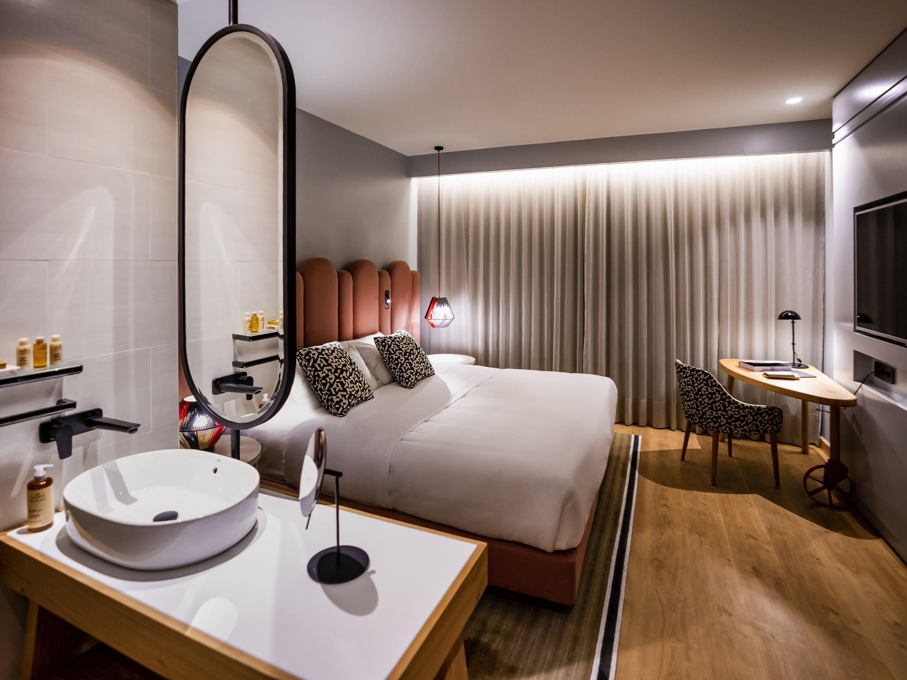 Habitación de hotel moderna con una cama grande, mesita de noche con lavabo blanco, espejo redondo, pequeño escritorio con lámpara y televisor.