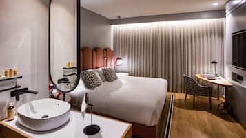 Habitación de hotel moderna con una cama grande, mesita de noche con lavabo blanco, espejo redondo, pequeño escritorio con lámpara y televisor.