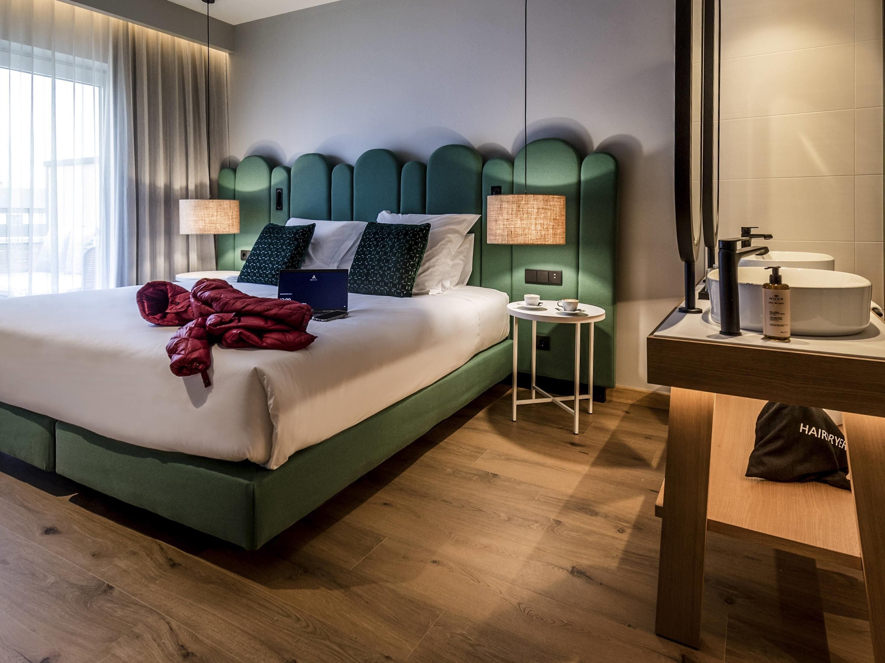 A hotel room with a bed, bedside lamps, a small table, a mirror, and a bathroom counter with toiletries.
