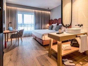 A hotel room with a bed, desk, chair, bedside table, mirror, and a view of the city. - Mercure Lugo Centro (Lugo)