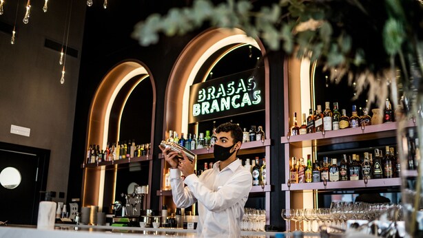 Un barman preparant begudes darrere d'una barra, amb amples prestatges plens de garrafes i gots.
