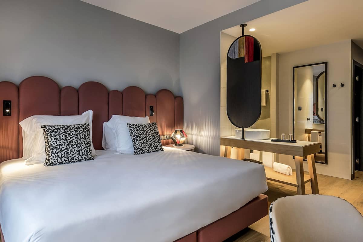 Un lit bien fait, avec une tête de lit, une table de chevet avec une lampe, un miroir et une chaise.