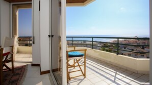 Een balkon met uitzicht op zee en gebouwen, een stoel met kussen, en een kamerkleed met patroon.