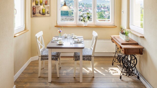 A dining area with a table set for four, two chairs, a window with a view of a town, a hanging light, and a small table with plants.