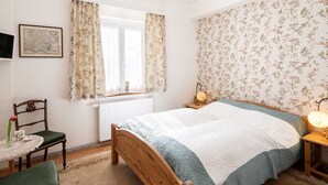 A bedroom with a wooden bed, floral wallpaper, a window with curtains, a bedside table with a lamp, and a chair with a table.