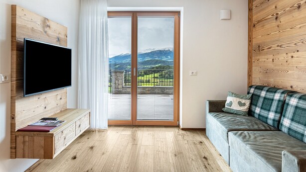 Een moderne woonkamer met een flatscreen televisie, houten paneelwanden, een grijze bank en een schuifdeur die leidt naar een balkon met uitzicht op bergen.