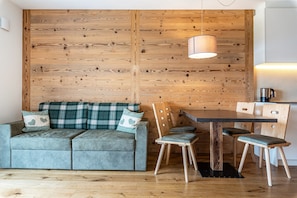 A cozy room with a wooden wall, a sofa with plaid cushions, a dining table with chairs, and a hanging light fixture.