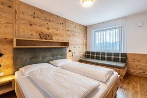 A bedroom with a wooden wall, a bed with white linens, a bench with a plaid cushion, and a window with sheer curtains.