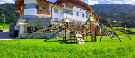 A two-story chalet with a play area, stone facade, and a mountain backdrop.