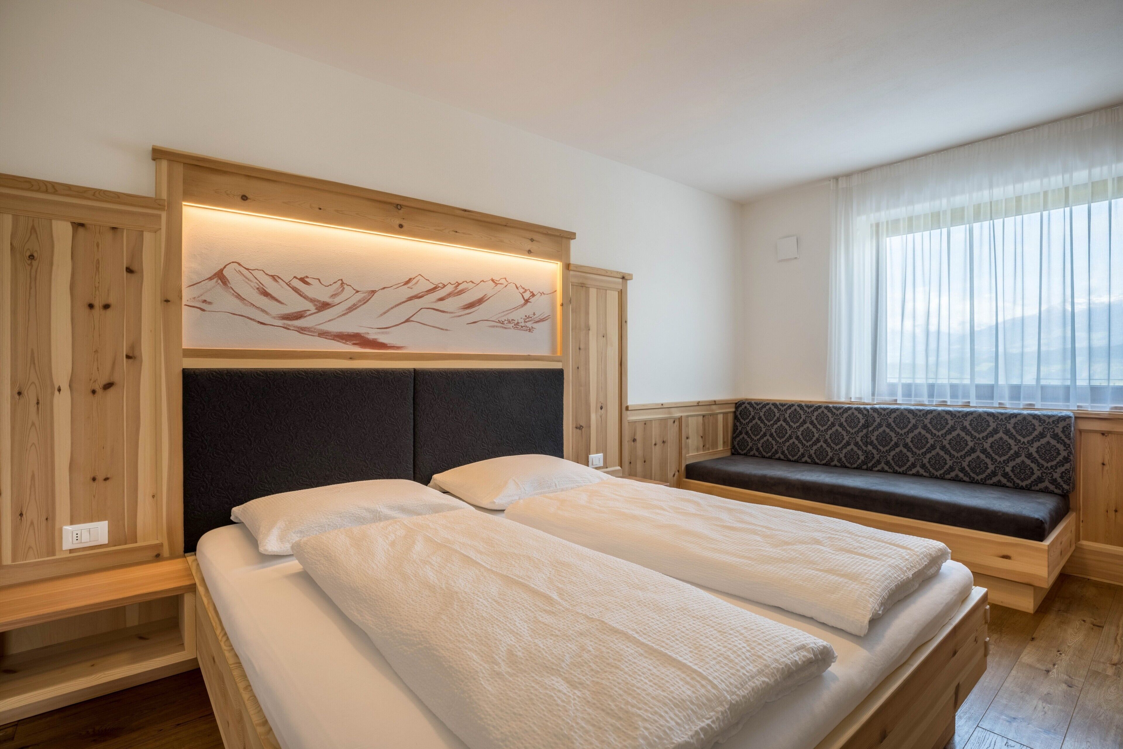 Ein Zimmer mit zwei Betten, einer Bank, einem Bergbild und einem Fenster mit durchsichtigen Vorhängen.