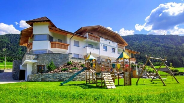 A two-story chalet with a play area, stone facade, and a mountain backdrop. - Apartment "Stefansdorf Wohnung 5" with Mountain View, Wi-Fi, Garden and Balcony (St. Lorenzen)