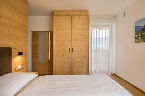 A bedroom with a wooden wardrobe, a bed with white bedding, a nightstand, a wall-mounted light, and a window with sheer curtains.