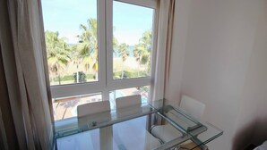 A room with a glass table and chairs, a large window with a view of palm trees and a beach. - El Amanecer 81 by homeprive (Denia)