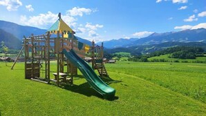 Un terrain de jeux avec un toboggan et des structures d’escalade, situé dans un champ herbeux avec des montagnes en arrière-plan.