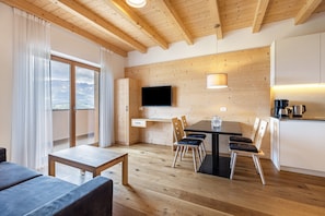 A modern kitchen with wooden cabinetry, a dining table with chairs, a flat-screen TV mounted on the wall, and a view of the mountains through large windows.