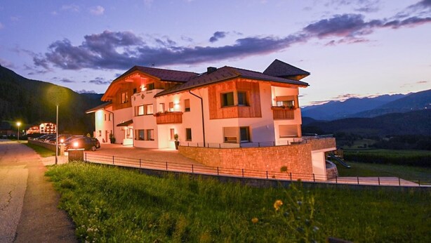 Un edificio de dos pisos con un balcón, una entrada para vehículos y el paisaje de montañas al fondo.